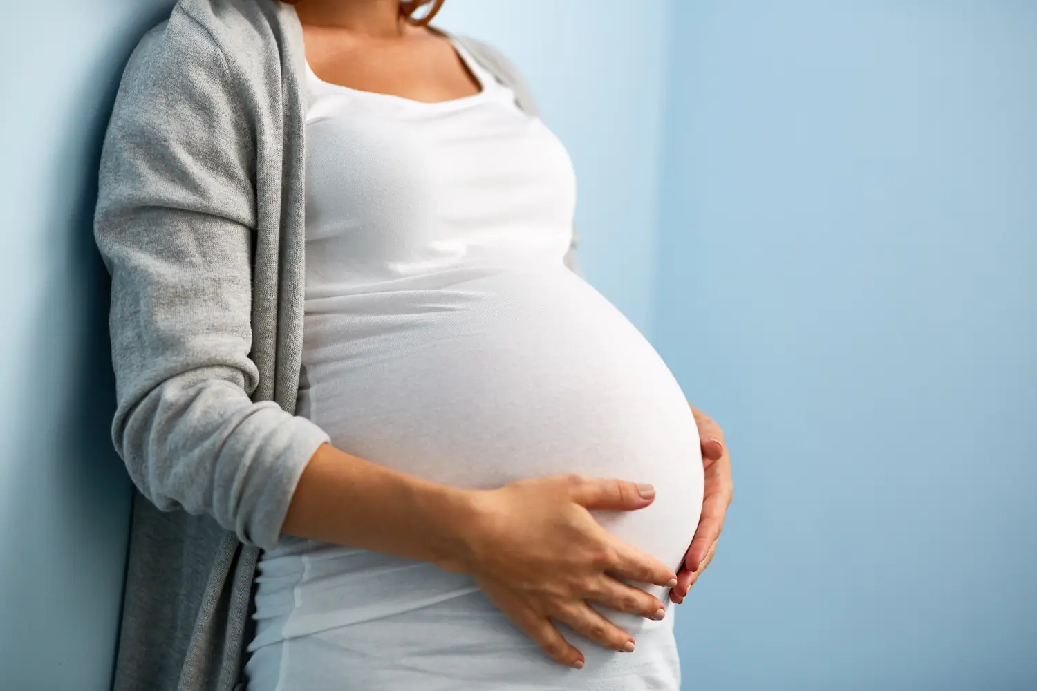Porträt einer nicht erkennbaren Frau in den letzten Monaten der Schwangerschaft, die ihren dicken Bauch sanft gegen die Wand in einem blauen Raum hält 